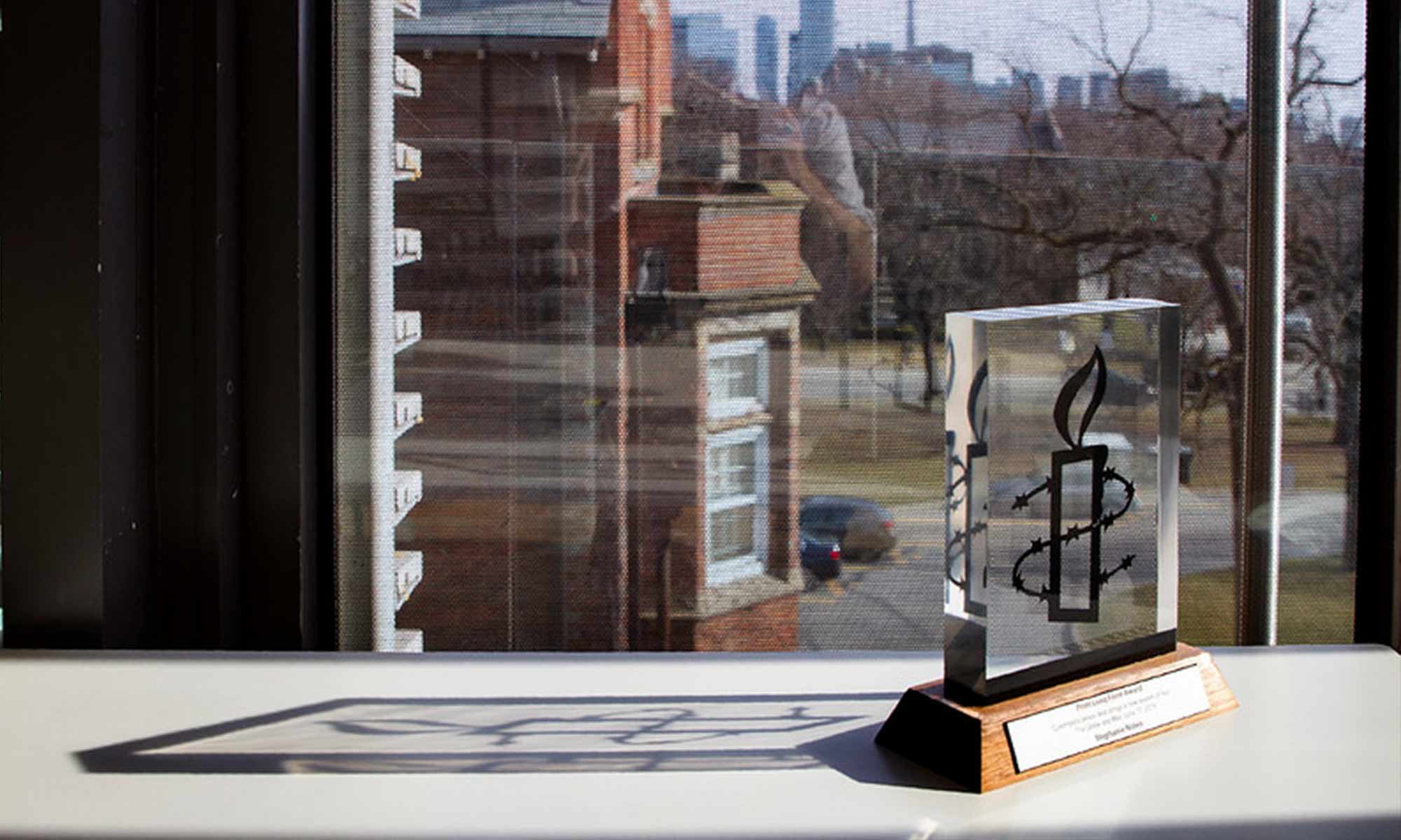 A square glass trophy with a wooden base sitting on a table in front of a window.