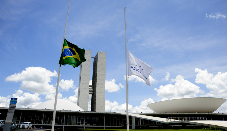 A green, blue and yellow Brazilian flag and a white Mercosur flag stand at half mast in front of a concrete building on a sunny day with partially cloudy skies.