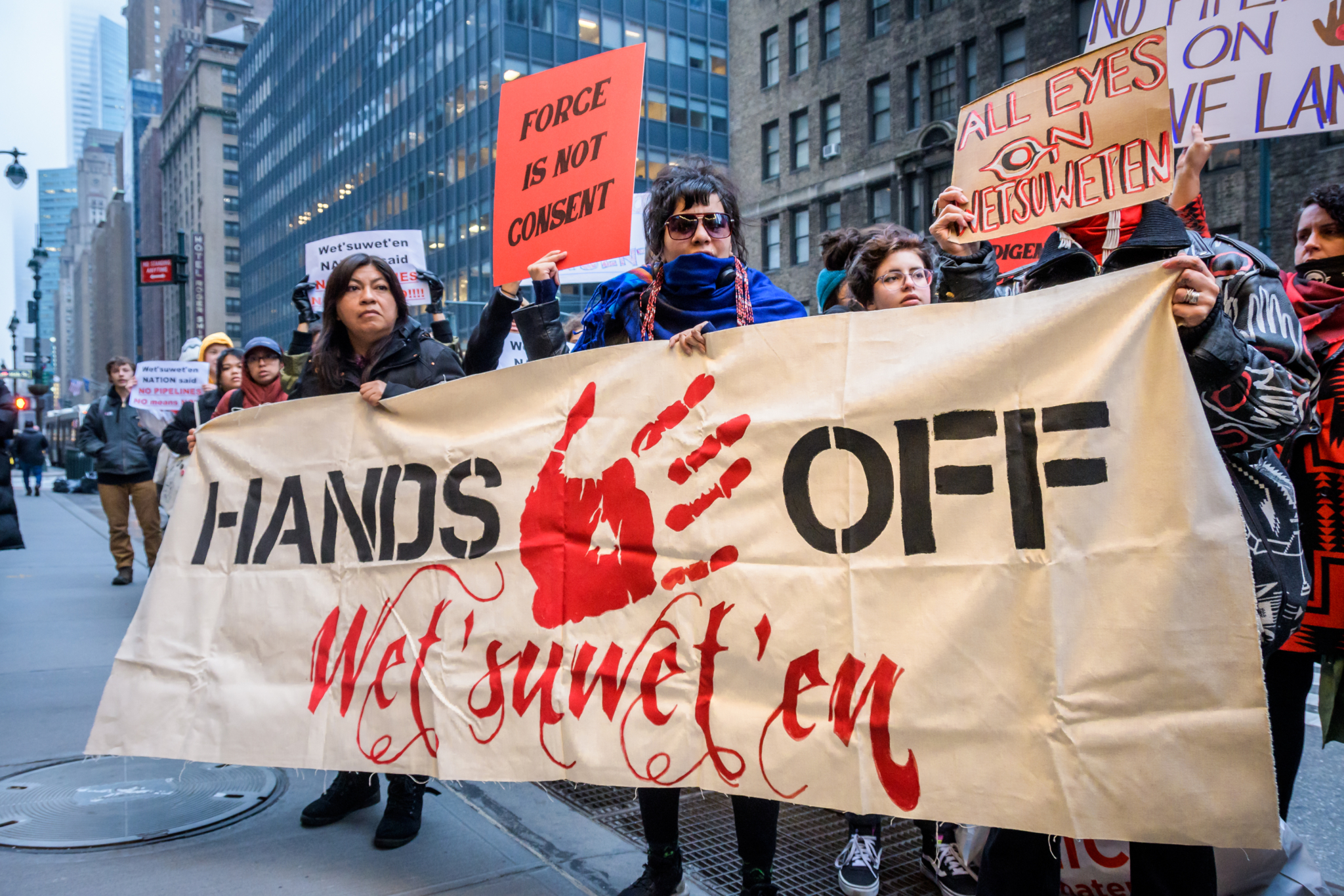 A group of protesters on the sidewalk hold up a banner stating "Hands Of Wet'suwet'en" and featuring a red hand print in the middle of the text.