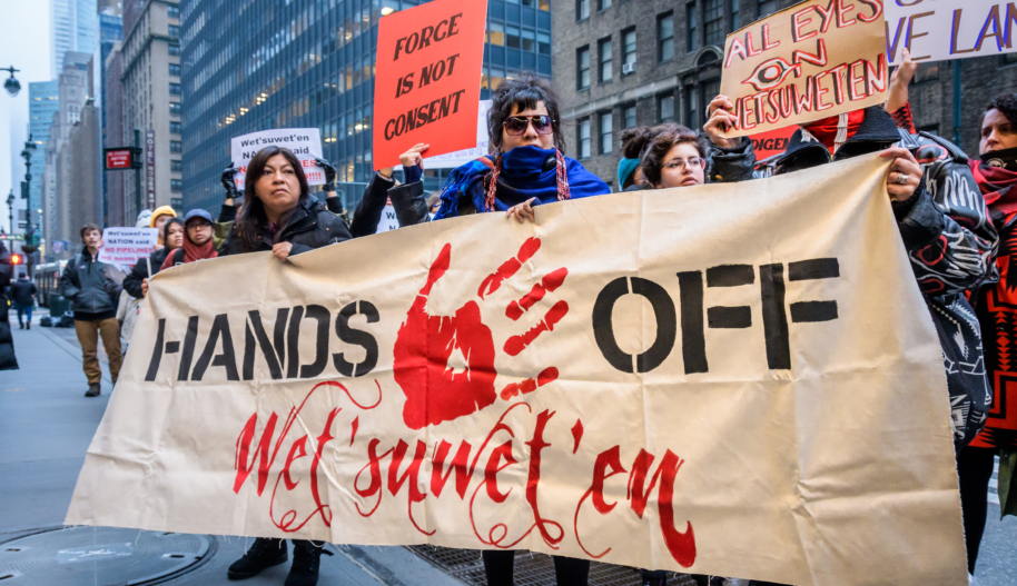 A group of protesters on the sidewalk hold up a banner stating "Hands Of Wet'suwet'en" and featuring a red hand print in the middle of the text.