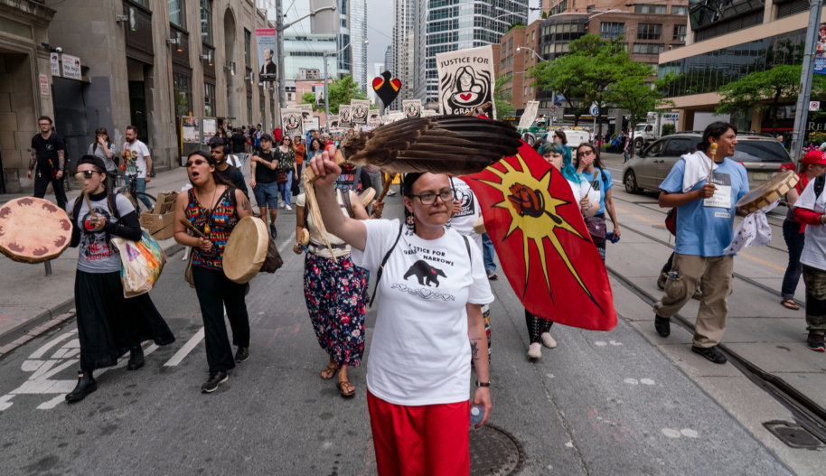 A woman wearing a white t-shirt and red pants holds a feather while she participates in a march for Indigenous rights on a street in downtown Toronto.