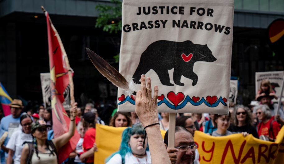 A hand holds up a feather at an Indigenous rights protest in front of a man holding a sign that says "Justice for Grassy Narrows."