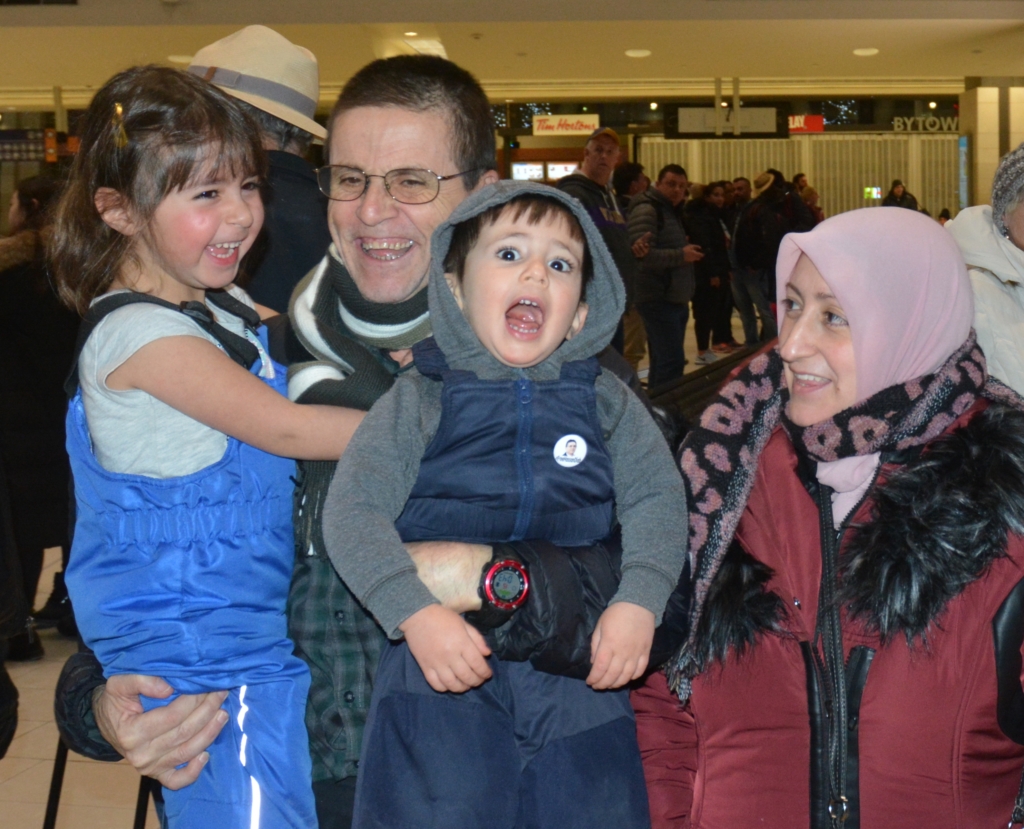 A smiling man holds his two young children, a boy and a girl, while his wife, also smiling, looks on.