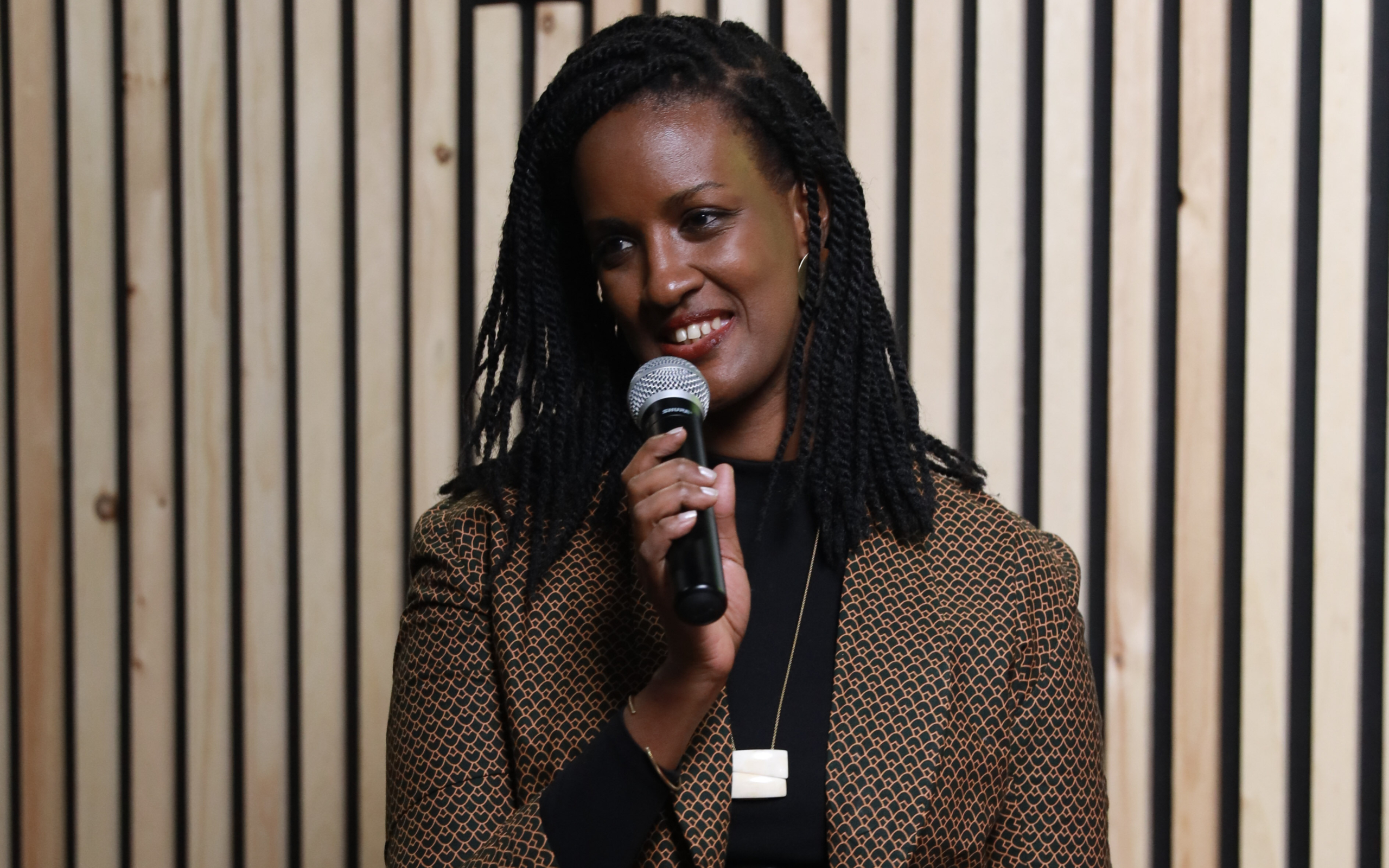 A woman in a tweed blazer and below-shoulder-length braids smiles while speaking into a microphone.
