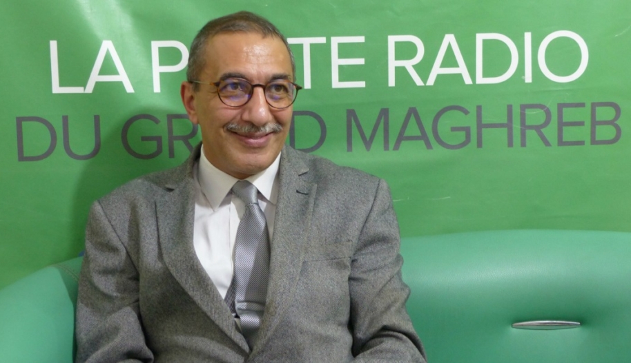 Ihsane El Kadi wearing a grey suit over a white shirt and grey tie, seating in front of a green poster with an inscription.