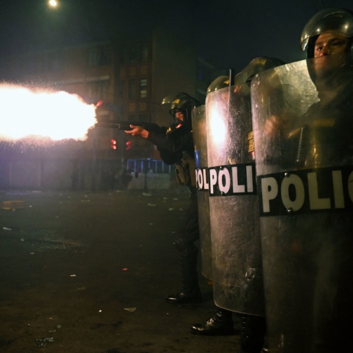 Two police officers in riot gear hold shields while another police officer behind them fires a gun.