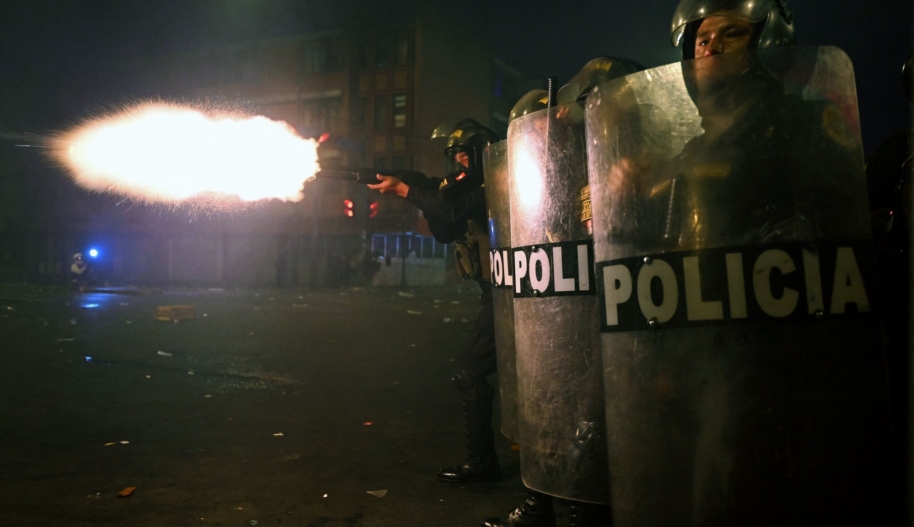 Two police officers in riot gear hold shields while another police officer behind them fires a gun.