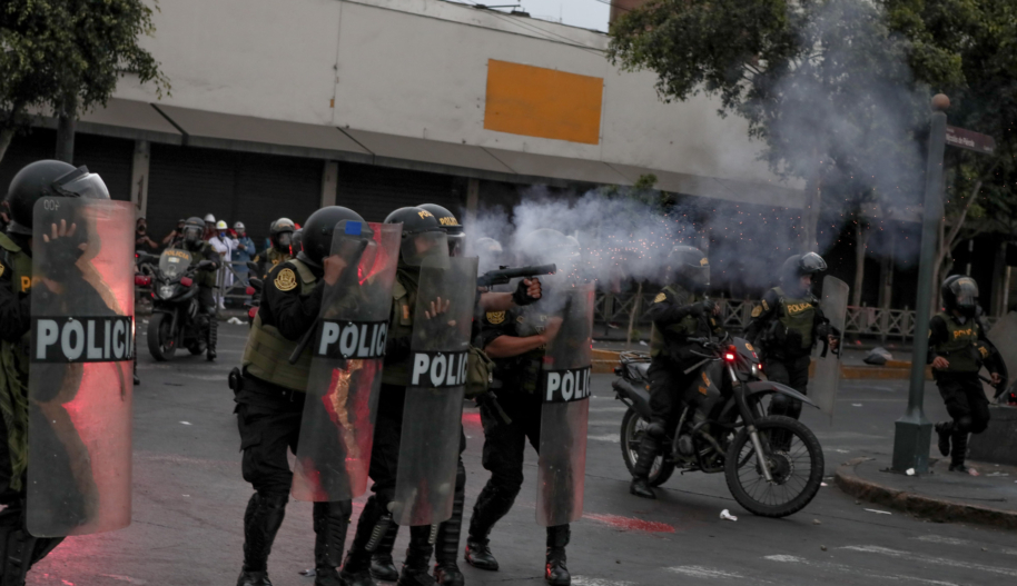 Four police in riot gear hold full-body-length shields and fire what looks like tear gas towards protesters