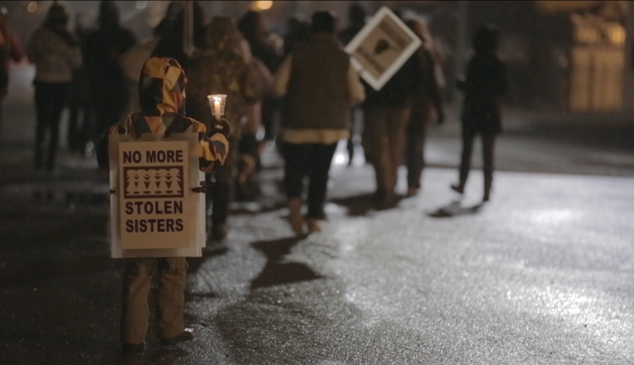A child holds a sign that reads "No More Stolen Sisters" at a rally on a road at night.