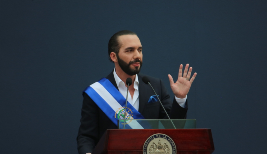 A man with a beard and slicked-back black hair speaks at a podium with his left hand raised in the air.